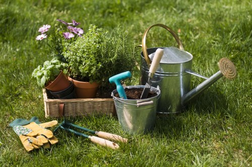 A garden scene featuring a lush, well-maintained lawn with vibrant green grass. In the foreground, there is a collection of gardening tools and accessories, including yellow gardening gloves, a pair of pruning shears, and small manual garden tools with wooden handles. To the left, a wicker basket holds potted plants and herbs with purple, green, and white foliage, adding colour and variety to the space. In the background, a silver metal watering can with a wooden handle rests on the grass, complemented by a small, round garden trowel attached to its side. The environment appears to be an outdoor backyard or side garden area, suggesting regular garden care activities typical of professional gardening services in Wandsworth, aiming to maintain healthy lawns and vibrant plant beds under natural weather conditions. The overall scene captures a moment of gardening preparation or tidying, with a focus on practical tools and lush plant displays typical of professional landscape maintenance in London.