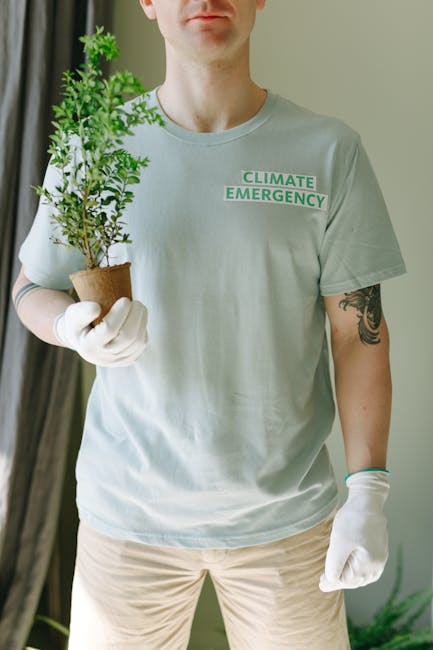 A person wearing a light grey T-shirt with the text 'CLIMATE EMERGENCY' and white gloves, is holding a small potted evergreen shrub with green, needle-like leaves. The individual has a visible tattoo 