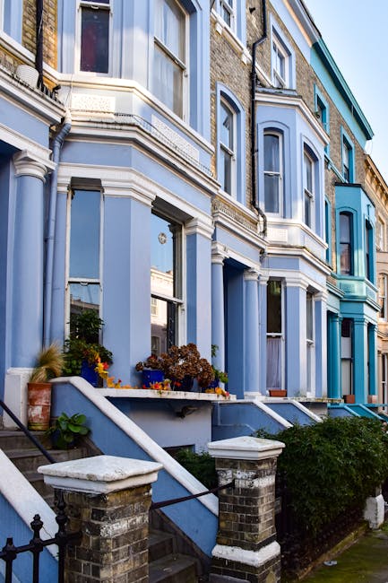 A row of Victorian terraced houses with ornate bay windows and decorative moldings, painted in varying shades of light blue and turquoise. The front gardens feature small steps leading up to the entra